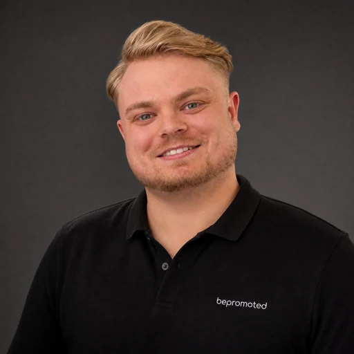 Tom Bach, blond mit kurzem Bart, im schwarzen bepromoted-Polo vor dunkelgrauem Hintergrund.